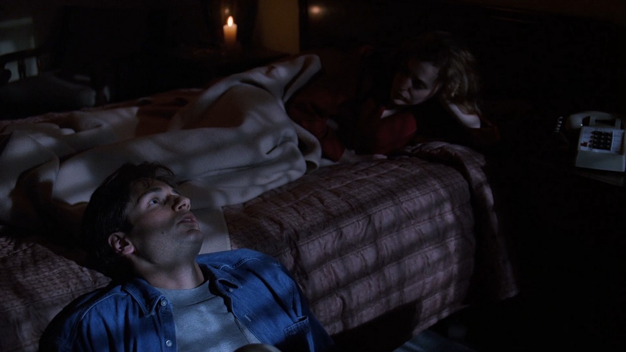 Mulder sitting beside the motel bed while Scully listens